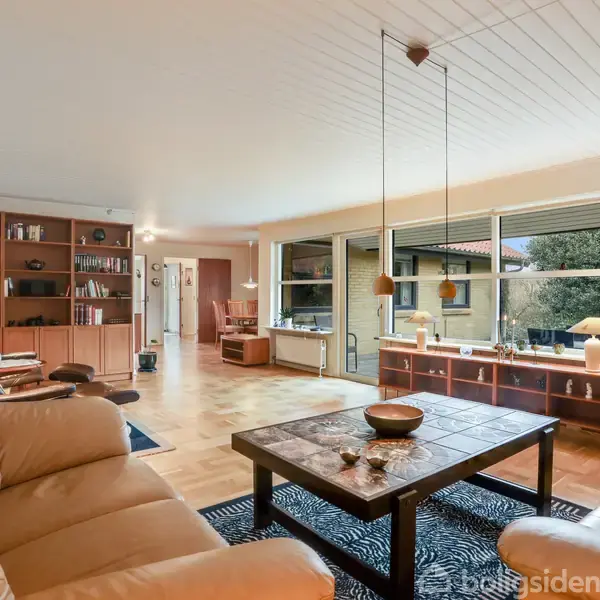 En lys stue med lyse lædersofaer om et kaffebord. En bogreol fylder væggen, store vinduer giver udsigt til en have, og lamper hænger over bordet.