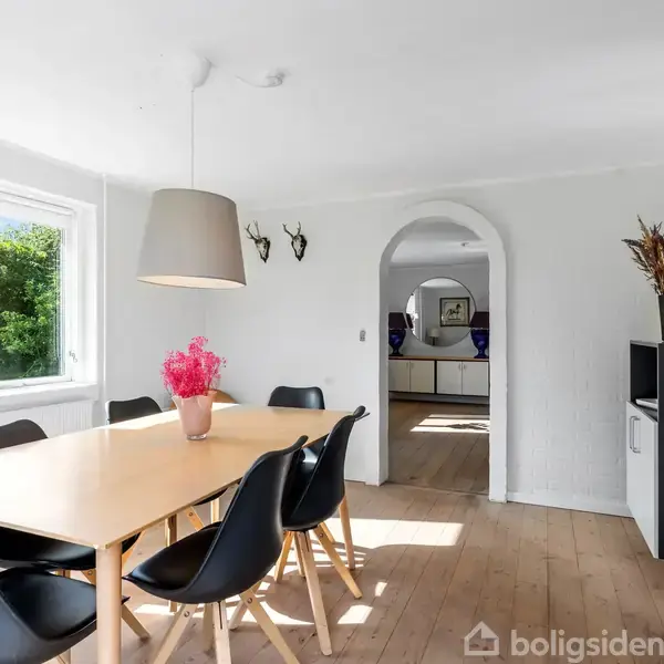 Spisebord omgivet af sorte stole står i et lyst rum med trægulv. En lampe hænger over bordet, og et vindue viser grønne træer udenfor. Blomster dekorerer bordet.
