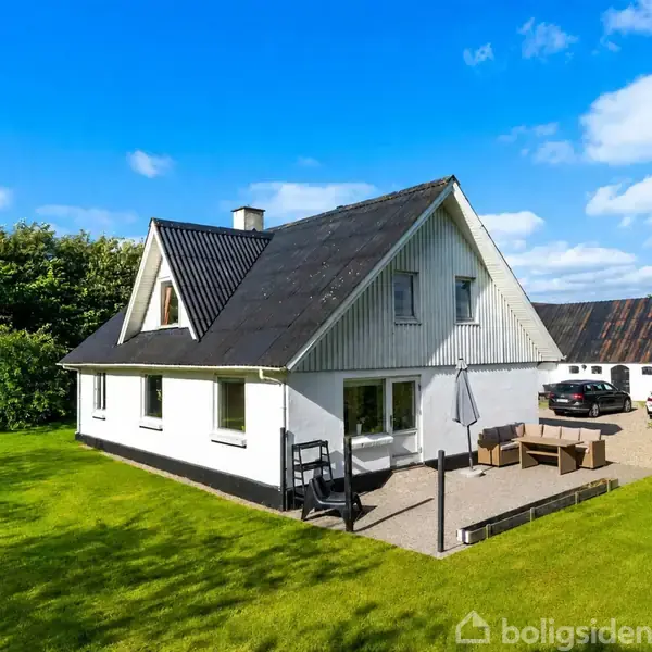 Et hvidt hus med sort tag står på en grøn græsplæne. Der er en terrasse med møbler og en grusindkørsel med parkerede biler. Skyfri blå himmel bagved.
