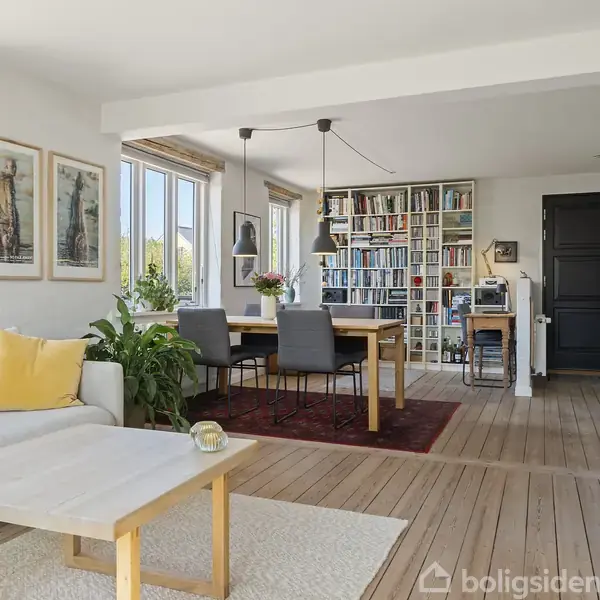 En stue med en hvid sofa og sofabord til venstre, der står ved siden af en spisebord med stole. Reoler fyldt med bøger fylder bagvæggen, belyst af store vinduer.