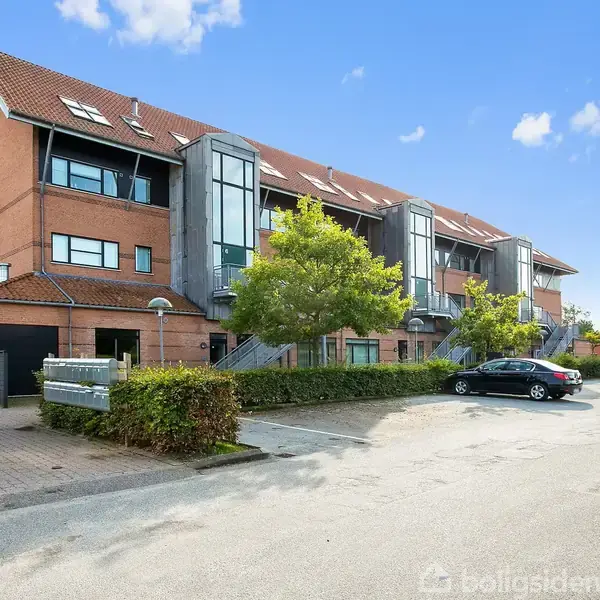 Bygningen er en moderne boligkompleks med flere etager og store glaspartier. Biler er parkeret udenfor i en asfalteret gård. Omkring bygningen er der grønne buske og træer under en blå himmel.