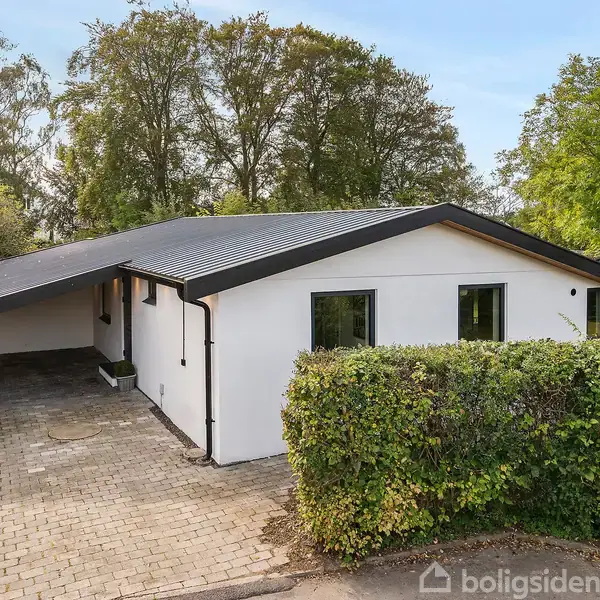 Et hvidt hus med fladt tag står stille ved en flisebelagt indkørsel omgivet af grønne hække og frodige træer på en solrig dag.