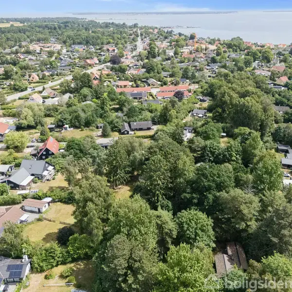Boligområde med huse omgivet af tæt skov og grønne områder, set fra oven; kystlinje og havet synligt i baggrunden.