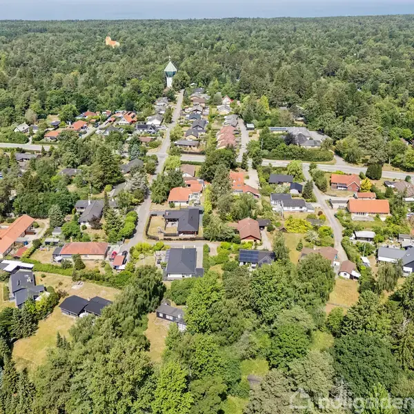 Landsbyområde set fra oven, med små huse spredt mellem grønne træer og buske. En vandtårn ses i baggrunden omgivet af skov. Det er en solrig dag.