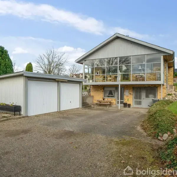 Et moderne hus med store glasvinduer står på en grusvej. Til venstre er en garagebygning med to porte. Græs og træer omgiver området på en klar dag.