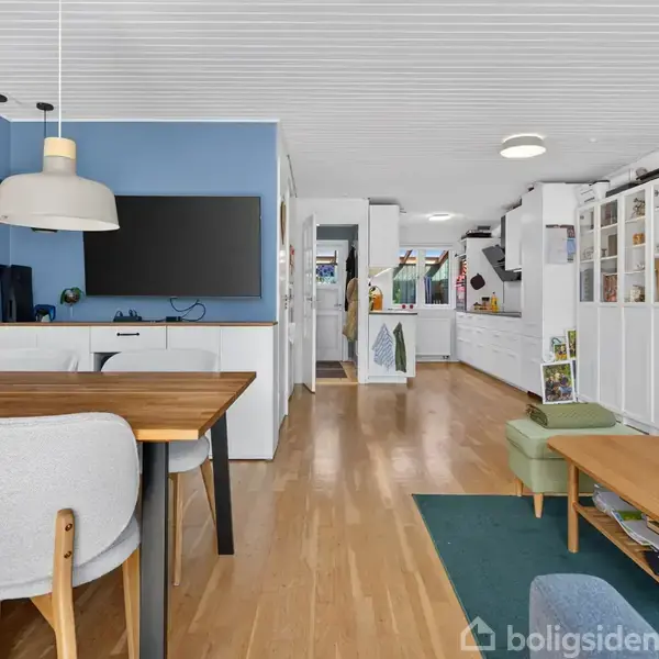 Et moderne stueområde med spisebord, stole og vægmonteret TV på blå væg; i baggrunden ses et køkken med hvide skabe. Gulvene er af lyst træ.