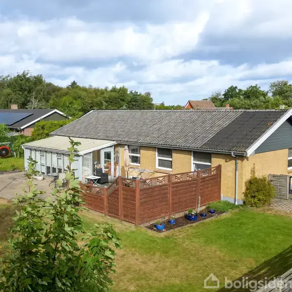 Et enfamiliehus med fladt tag og gult murværk står stille omgivet af en grøn have med græsplæne og terrasse indrammet af træhegn i et villakvarter.