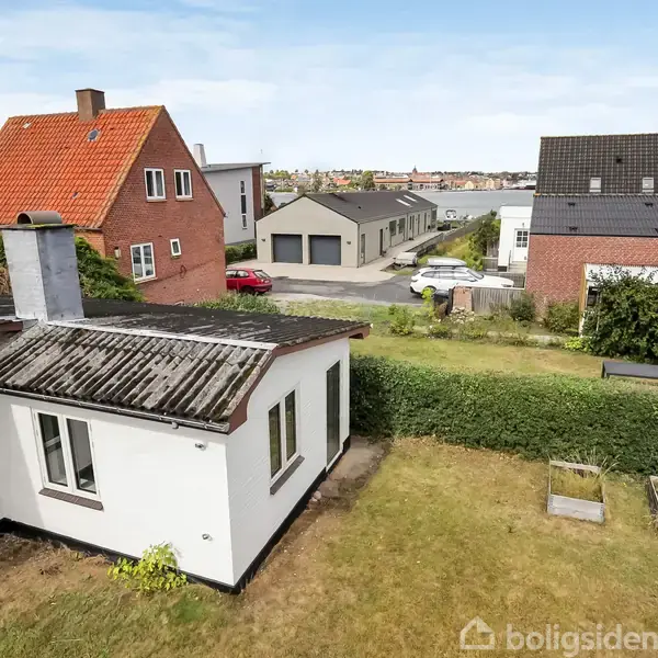 Huset står stille i en forstadshave, omgivet af græs og buske. Flere huse ses i baggrunden, med en blå himmel over dem.