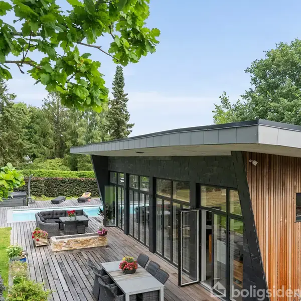 Moderne træhus med fladt tag, omgivet af træer. Store vinduespartier åbner ud mod en terrasse med siddepladser og en pool. Grøn have omgiver området.