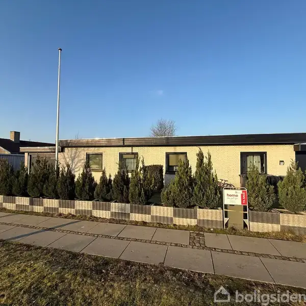 En enkelt-etagers gult murstenshus står bag en række stedsegrønne buske. Ved fortovet er et skilt fra "home". Blå himmel og solbelyst scene omkranser boligen.