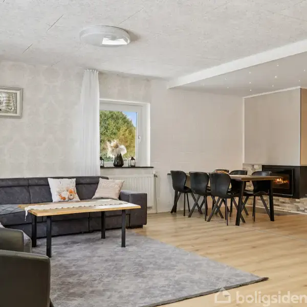 L-formet sofa står på gråt tæppe i stue, ved siden af spisebord med sorte stole. Pejsen indbygget i væggen. Vindue viser udsigt til træ og buske.