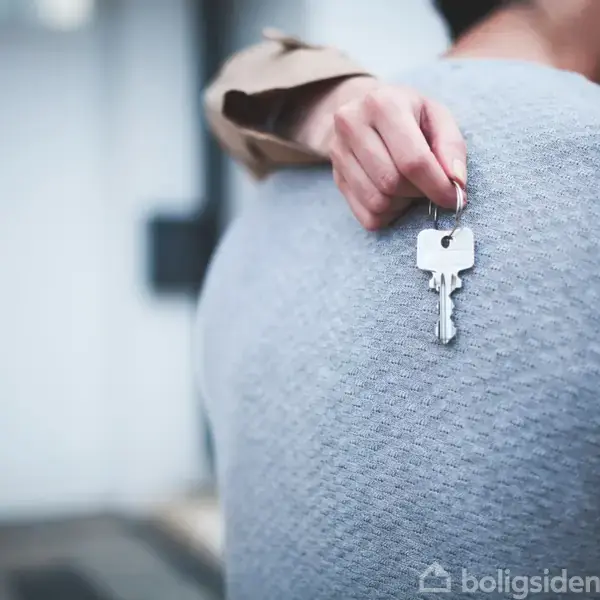 En person krammer en anden, mens vedkommende holder en nøgle i hånden. De står foran en uklar dør, hvilket indikerer en boligkontekst.