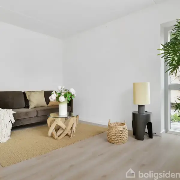 En brun sofa med puder og et tæppe står i en lys stue. Foran sofaen er et rundt træbord med blomster. En stor plante og en lampe står ved et vindue.