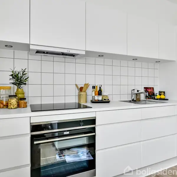 Køkkenområde med hvidt interiør. Komfur og ovn er centreret under et hvidt flisespejl. Bordpladen er dekoreret med skærebrætter, planter og krydderier. Rummelig og minimalistisk atmosfære.