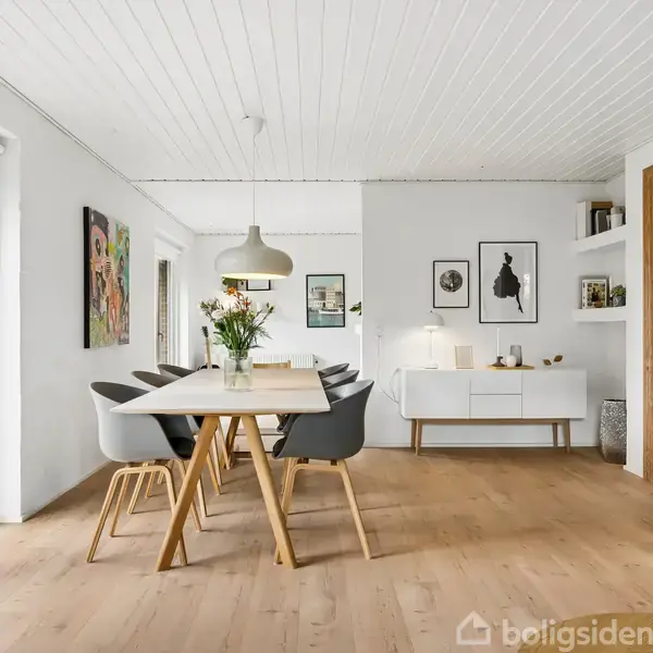 Spisebord med blomster i vase på bordet står i moderne, lyst indrettet spisestue med træmøbler og kunst på væggene. Store vinduer til venstre giver udsigt til en terrasse.