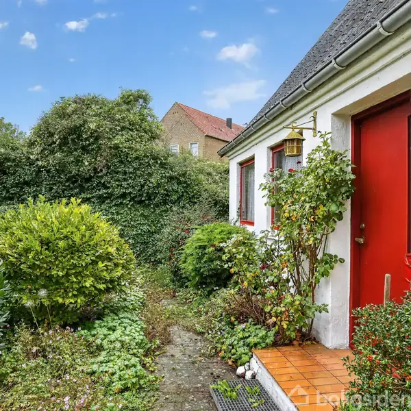 Huset har en rød dør og nummer 15 på væggen. Omgivet af en frodig have med buske og planter, under en blå himmel.