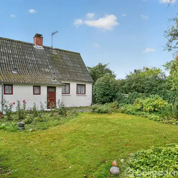 Hvidt murstenshus står stille omgivet af en velplejet have med græsplæne og buske, under en blå himmel med spredte skyer.