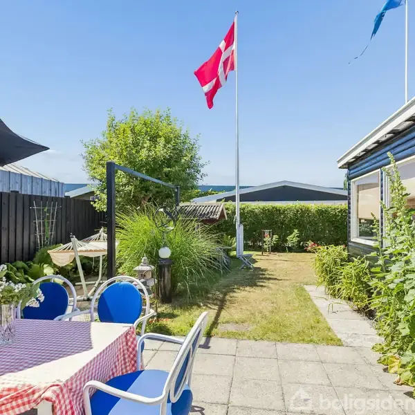 Et bord med blå stole står på en terrasse med rødternet dug. Et dannebrog blafrer i haven ved siden af et sort hus og grønne buske under en klar blå himmel.