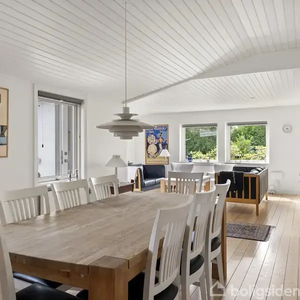 En spisestue, med et langt træbord og hvide stole, oplyst af en hængelampe, fører til en stue med store vinduer og grøn udsigt. Plakater: "MØN" og en reklameplakat.