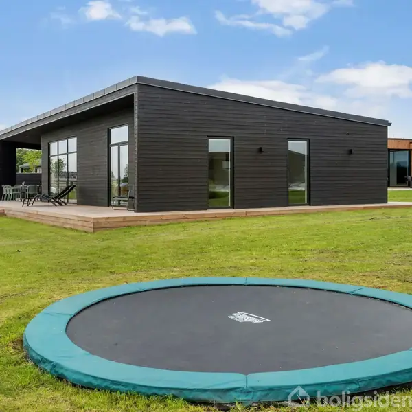 En moderne, sort træbygning står på en græsplæne. Der er store glasdøre og vinduer. Foran ses en nedgravet trampolin i græsset. Solrigt vejr med blå himmel og få skyer.