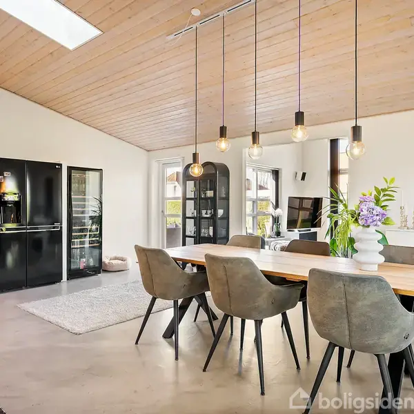 Et spisebord med otte stole står i en moderne stue med trægulv og loft. Loftslamper hænger over bordet. Rummet har et TV og hvid reol.
