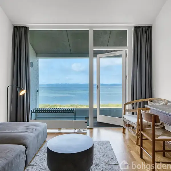 En græsplæne strækker sig mod stranden med en lang træbro ud i vandet. I baggrunden ligger en stor sejlskib ved en havn. Klar blå himmel.