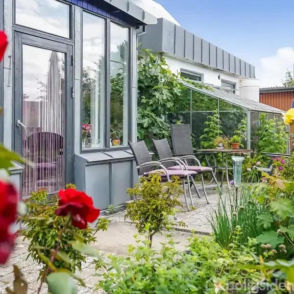 Glaspavillon med havemøbler står på terrasse omgivet af blomstrende roser og grønne planter. Parasollen er lukket, og himlen er delvist overskyet.