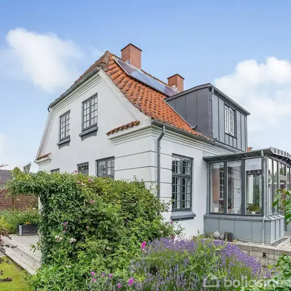 Hus med klassisk arkitektur har et rødt tegltag og hvide facader. Beliggende i en have med grønne planter og blomster, er det omkranset af en klar, blå himmel.