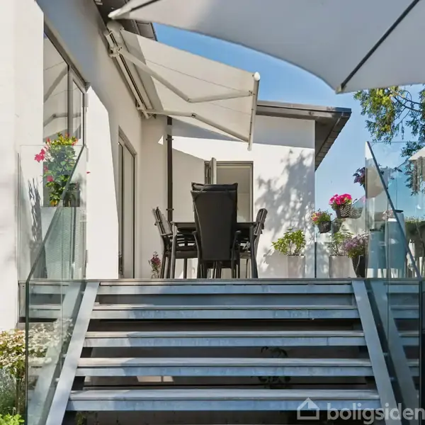 En terrasse med bord og stole er omgivet af glasrækværk. En parasol og markise giver skygge. Blomsterkrukker pynter området. Trapper fører op til terrassen fra en grøn have.