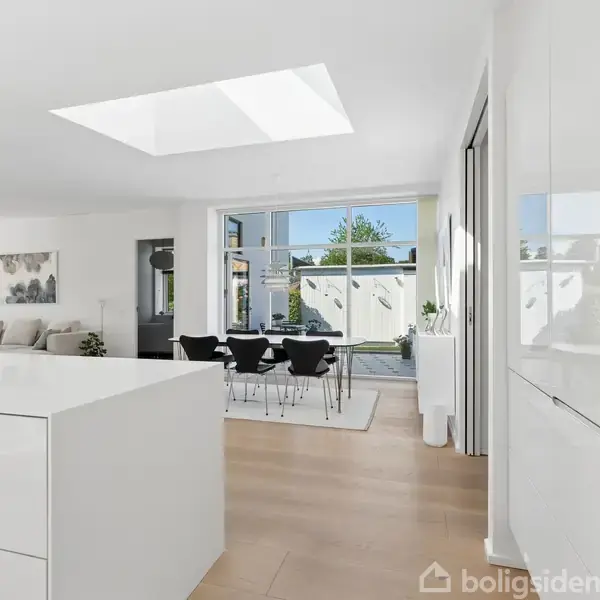 Et moderne køkken i hvidt design med en ø, lyser gennem ovenlys, åbner op til en spisestue med glasdøre. Trægulve og minimalistisk indretning dominerer rummet.