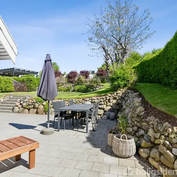 Terrassemøbler står omkring et bord under en parasol på en flisebelagt terrasse. Omkranset af stensætninger med grønne buske og træer, der skaber en privat og idyllisk haveomgivelser.