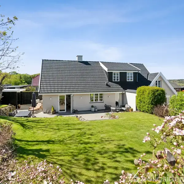 Et hus med sorte tagsten står i en have med grøn græsplæne. Omkring huset er der buske og træer. Himlen er klar og blå i baggrunden.