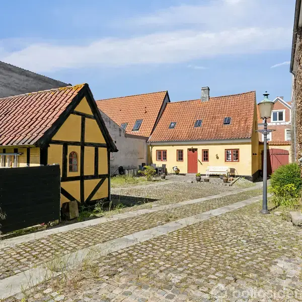 Et bindingsværkshus med rød tag. Husets facade er gul med sorte bjælker. Det står på en brostensbelagt gårdsplads omgivet af andre bygninger. Blåt himmel og solskin.