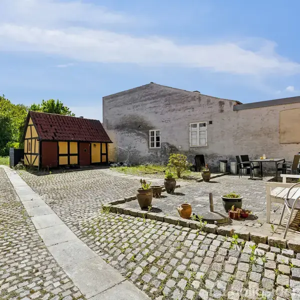 En lille bindingsværksskur står på en brolagt gårdsplads ved siden af en terrasse med havemøbler og krukker. Omgivet af vægge og grønne træer under en klar blå himmel.