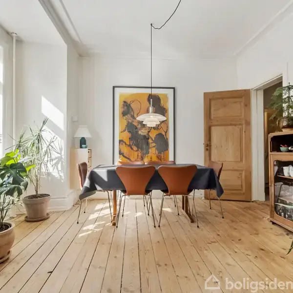 Et spisebord med en sort dug er omgivet af fire brune stole. Bordet står i et lyst rum med trægulv, planter og kunst på væggen. En åben dør er i baggrunden.