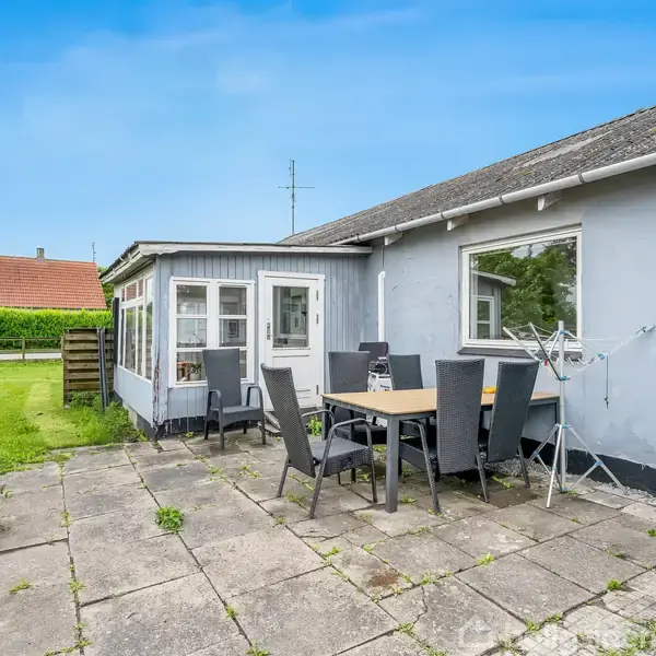 Et havebord med stole står på en flisebelagt terrasse ved siden af et gråt hus med store vinduer. I baggrunden ses en grøn græsplæne og et træhegn.