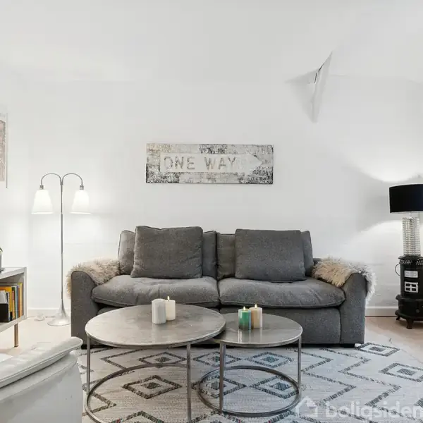 Grå sofa står centralt mod en hvid væg med "ONE WAY" skilt. Ved siden af to borde med lys. Rum med lys bordlampe, tæppe og vindue.