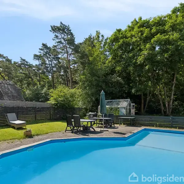 Swimmingpool omgivet af havemøbler på en terrasse ved en have. Træer og et stråtækt hus er synlige i baggrunden under en klar blå himmel.