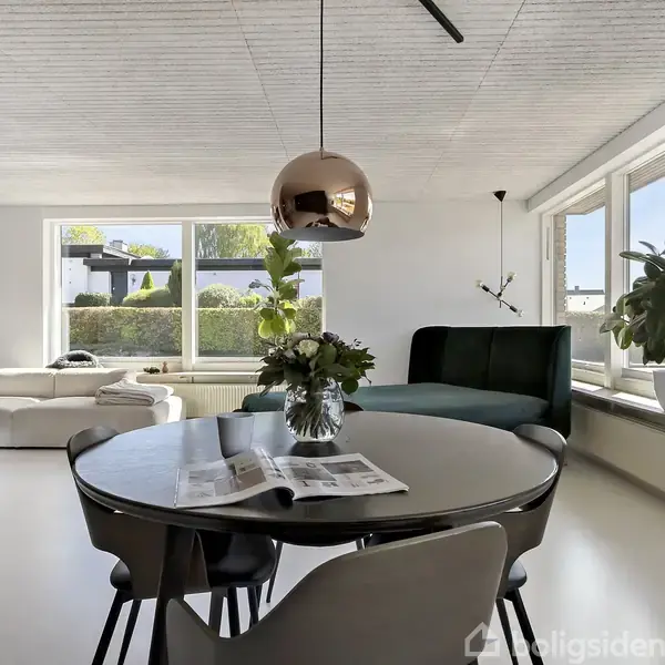 Rundt spisebord med blomster og avis står i et lyst opholdsområde, der har store vinduer, sofa og flere planter. Moderne, minimalistisk stil præger rummets indretning.