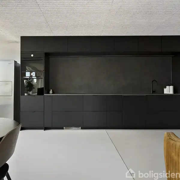 Et moderne køkken med sorte skabe. En indbygget ovn og et stort køleskab står til venstre. Et rundt bord med stol til venstre og en gul stol til højre.