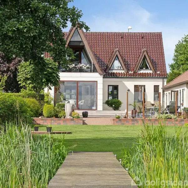 Et hus med rødt tegltag og trekantede vinduer står i en have med græsplæne og terrasse. En træsti fører gennem et bed med høje planter mod huset.