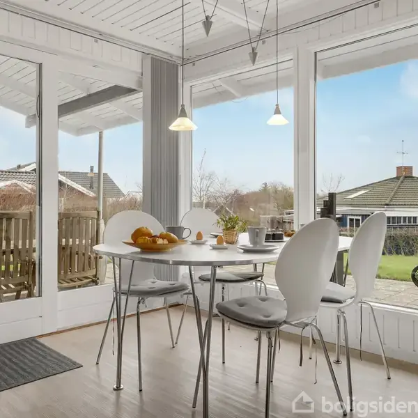 Et rundt spisebord med hvide stole er dækket til morgenmad med brød og drikkevarer. Det står i en lys udestue med store vinduer, der giver udsigt til en have.