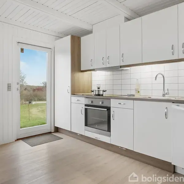 Køkken med hvide skabe, kogeplader, ovn og opvaskemaskine; står i et lyst rum med trægulve og udsigt til have gennem glasdør; vægdekoration til venstre.
