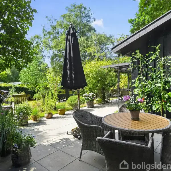 En rund træbord med flettede stole står på en gårdhave. Planter og blomster omgiver området, og en stor parasol tilbyder skygge. Baggrunden består af tæt, grøn vegetation.