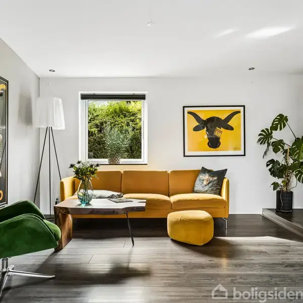 En stue med en grøn lænestol og en gul sofa, placeret ved et lavt sofabord. Vægge er dekoreret med plakater. Store vinduer slipper lys ind, og der er grønne planter.