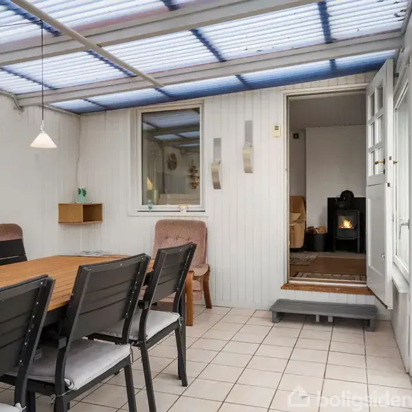 En terrasse med spisebord og stole står på flisegulv. En åbning fører til et indendørs rum med brændeovn. Lyse vægge og gennemsigtigt tag omfatter rummet.