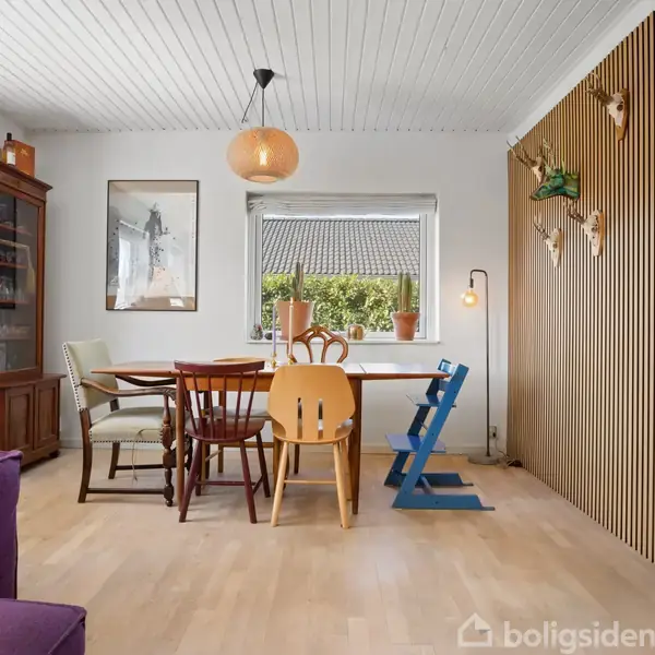 Spisebord med forskellige stole står i hjørne af lyst rum. Lampe hænger over bordet. Væg med trælister har dekorationer. Vindue viser tag og træer udenfor.