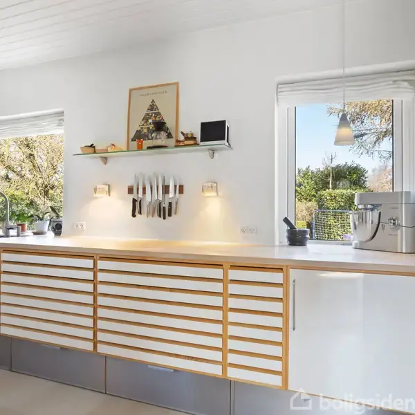 Køkkenbord med trælåger er centralt, hvor knive hænger på væggen. To vinduer med udsigt til have. Dekorationer og apparater som kaffemaskine og vase er på bordet.