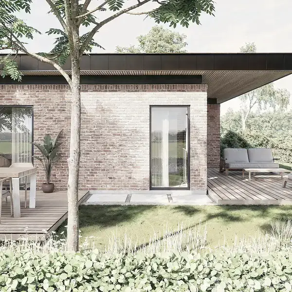 En moderne, enetages murstensvilla med store vinduer står på en træterrasse. Der er havemøbler med stole og en sofa, omgivet af grønne planter og buske, under klare himmelstræk.