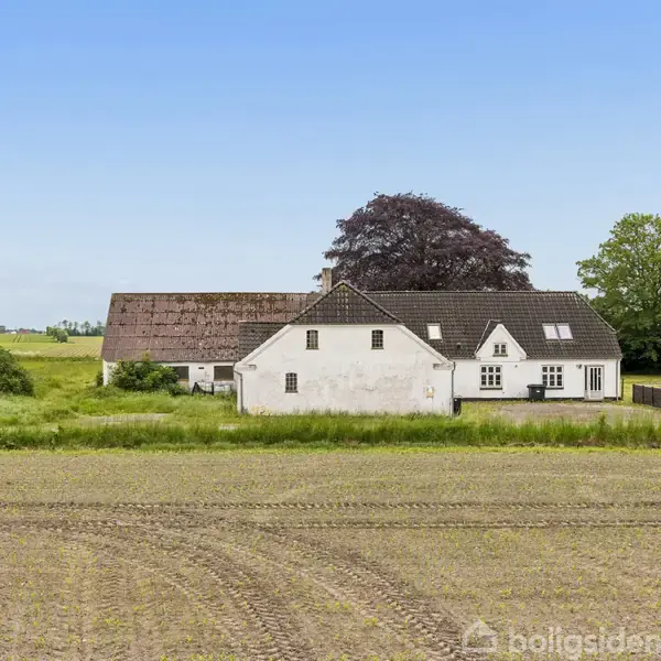 En hvid landbygning står stille foran en stor lade med et brunligt tag. Bygningerne er omgivet af marker og grønne træer under en klar blå himmel.